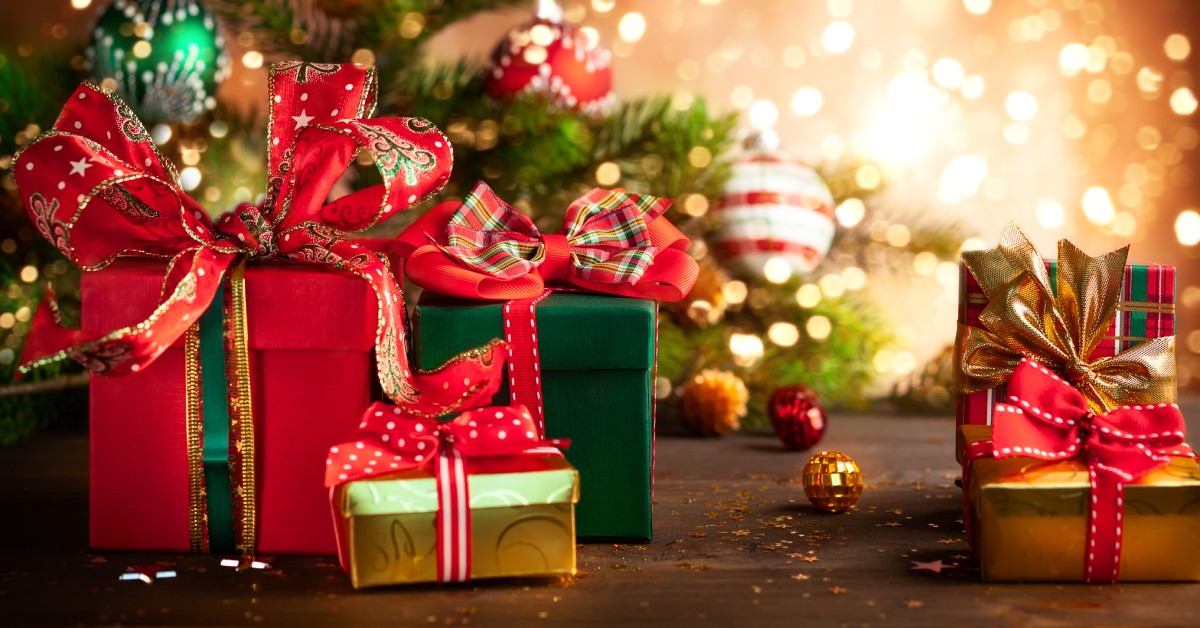 A ground view of Christmas gifts and decorations on a hardwood floor. Gifts include red, green, and gold boxes with ribbons.