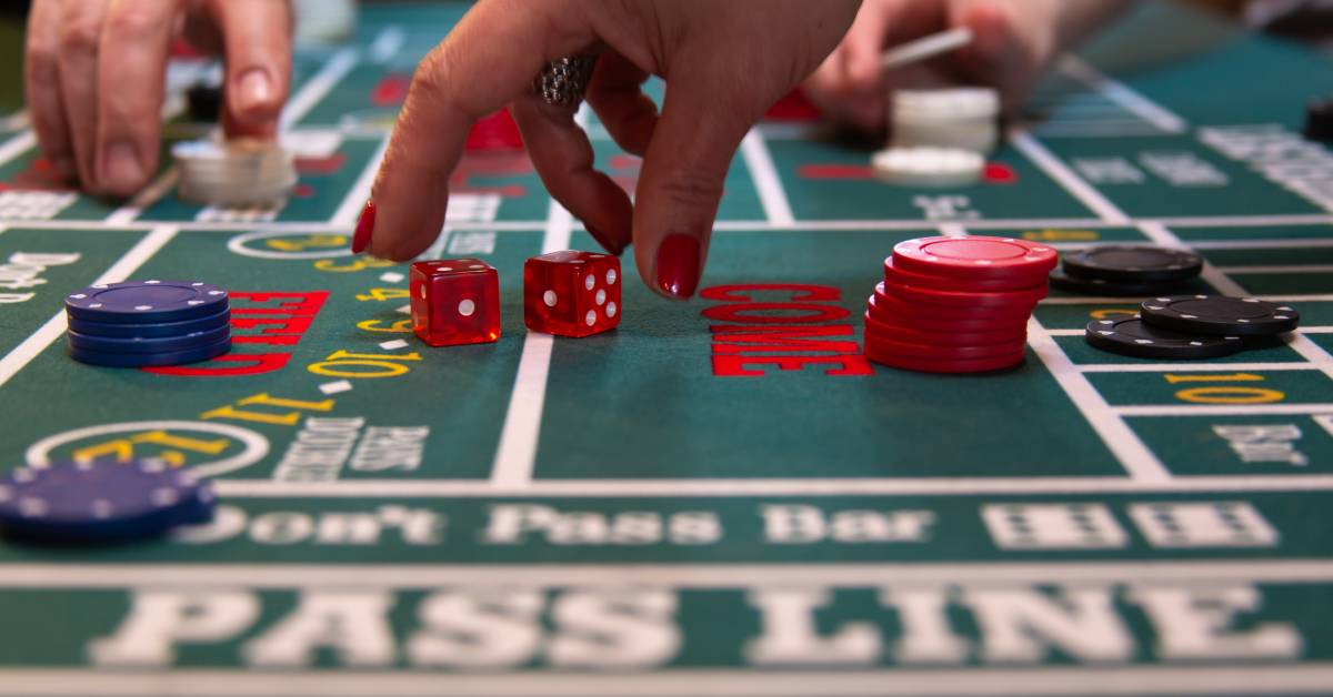 A woman with red painted fingernails picking up two red craps dice off a green craps game table felt.