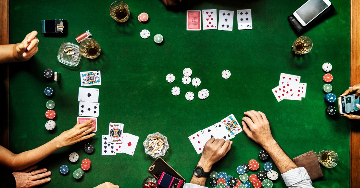 An overhead view of a group of people playing poker on a green felt poker table with cards, chips, and drinks.